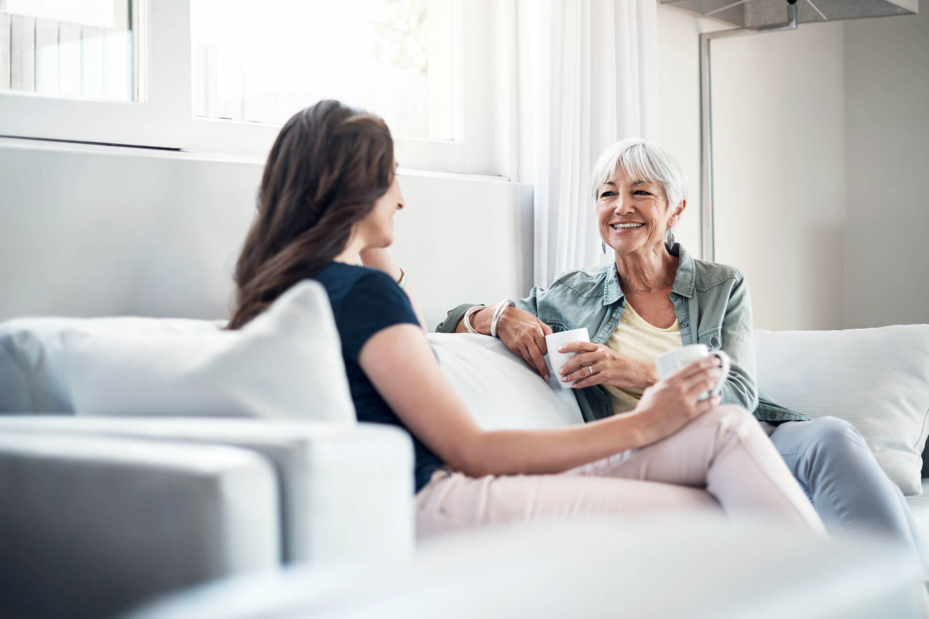 Two women sitting in a light room on a couch smiling while talking and holding coffee