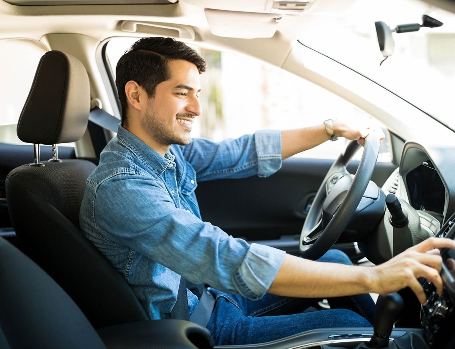 A man seated in a car adjusts the controls on the center console while smiling. He is wearing a denim shirt and appears relaxed.